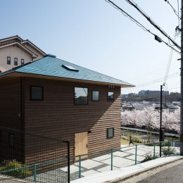 四つ角の家｜４つの小屋と立体路地の家｜大阪府堺市｜新築一戸建て住宅 (北西から見る。桜並木の先はなだらかな丘陵。アプローチは建物のかたちを踏襲した正方形のコンクリート土間。)
